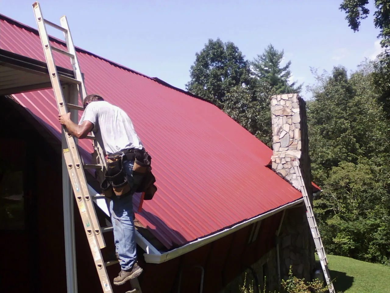 Roofer on a ladder beside a red metal roof and stone chimney — Soffit & Fascia Repair by Roofing Brothers in Erwinna